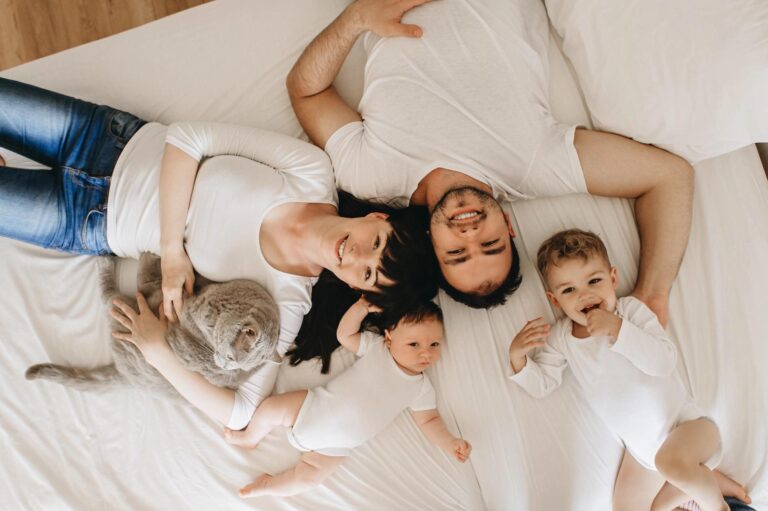 Happy family with mother, father, baby, toddler, and pet cat lying on a bright, clean white bed, symbolizing a safe and healthy home environment.